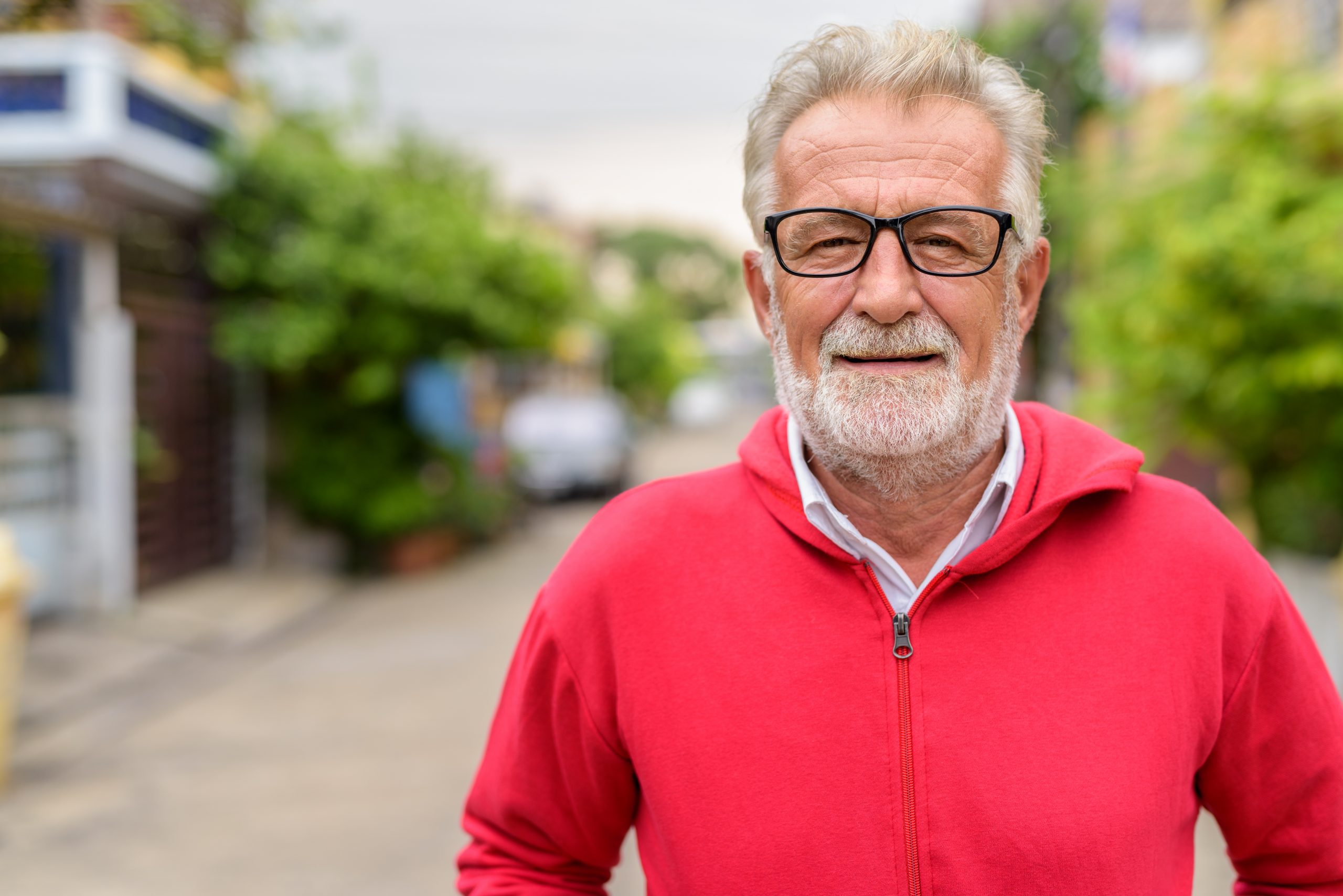 Happy handsome senior bearded man smiling while wearing eyeglasses and red jacket outdoors