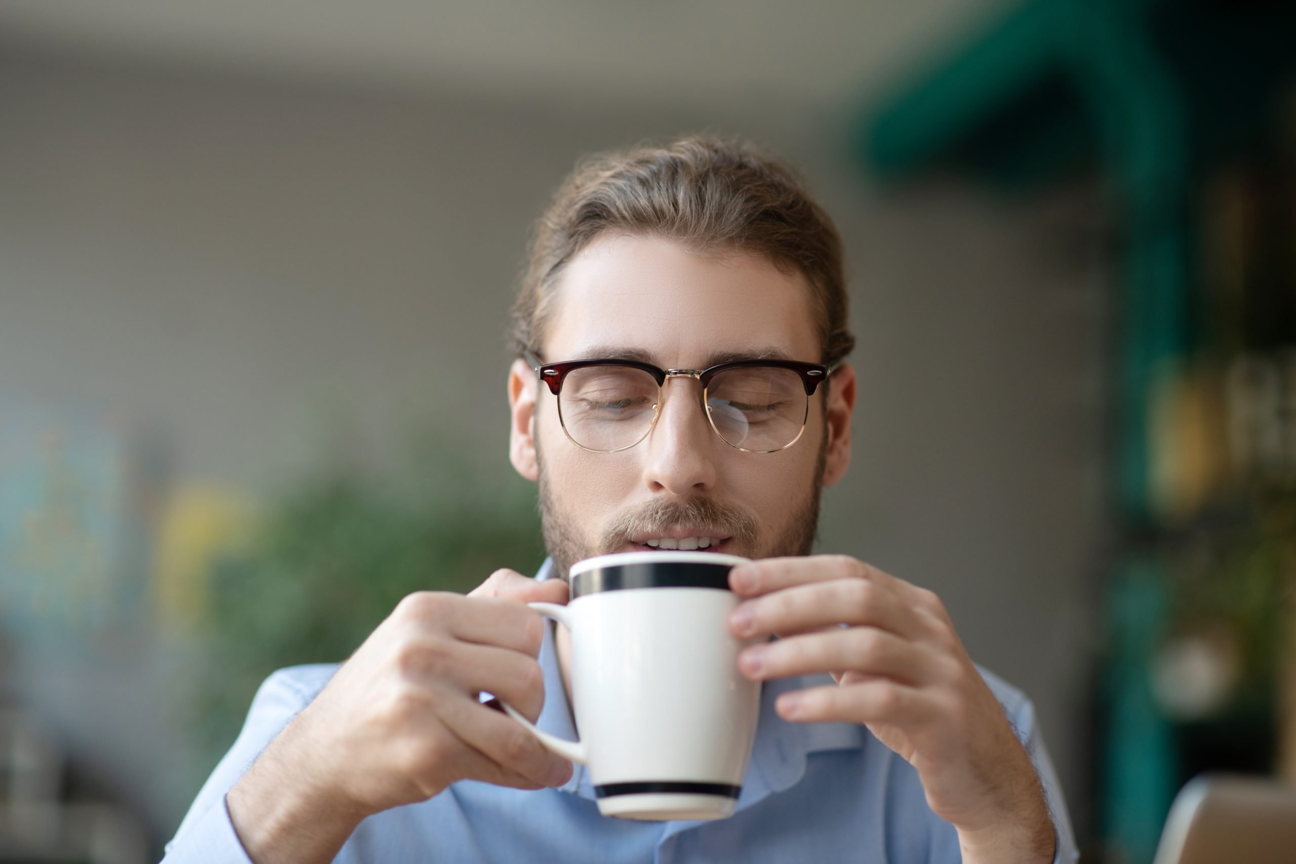 Time to have some coffee. Young joyful man with glasses looking smiling at a white cup with coffee that he holds in his hands, in good mood.